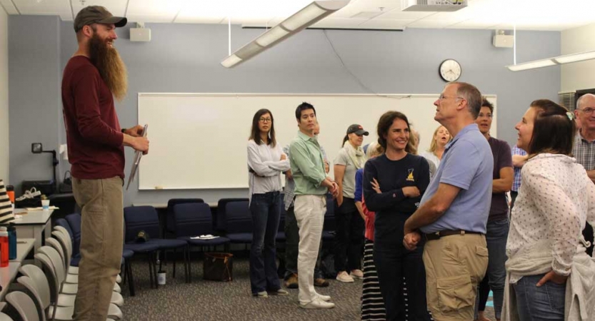 A person stands on a chair, speaking to a group of people at the family seminar of an outward bound intercept course